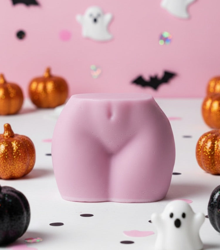 Pink buttocks-shaped candle on a Halloween-themed background with pumpkins and ghosts.